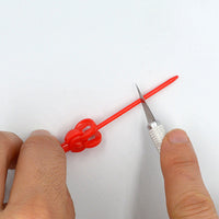 Hands demonstrating how to split AK Interactive holding pin along the pre-scored groove using a hobby knife blade showing the red plastic clip-top pin being separated into two halves