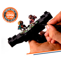 Hand holding AK Interactive Painting Handle XL platform while painting a multi-figure diorama scene with two colourful fantasy characters on a scenic base, demonstrating the wide grip platform for large bases with 40mm and larger badge