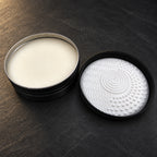 Open tin of cream solid brush soap next to its lid with the white silicone cleaning pad, seen from above