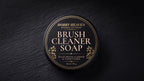 Closed black screw-top tin with the gold-foil Brush Cleaner Soap label on dark slate