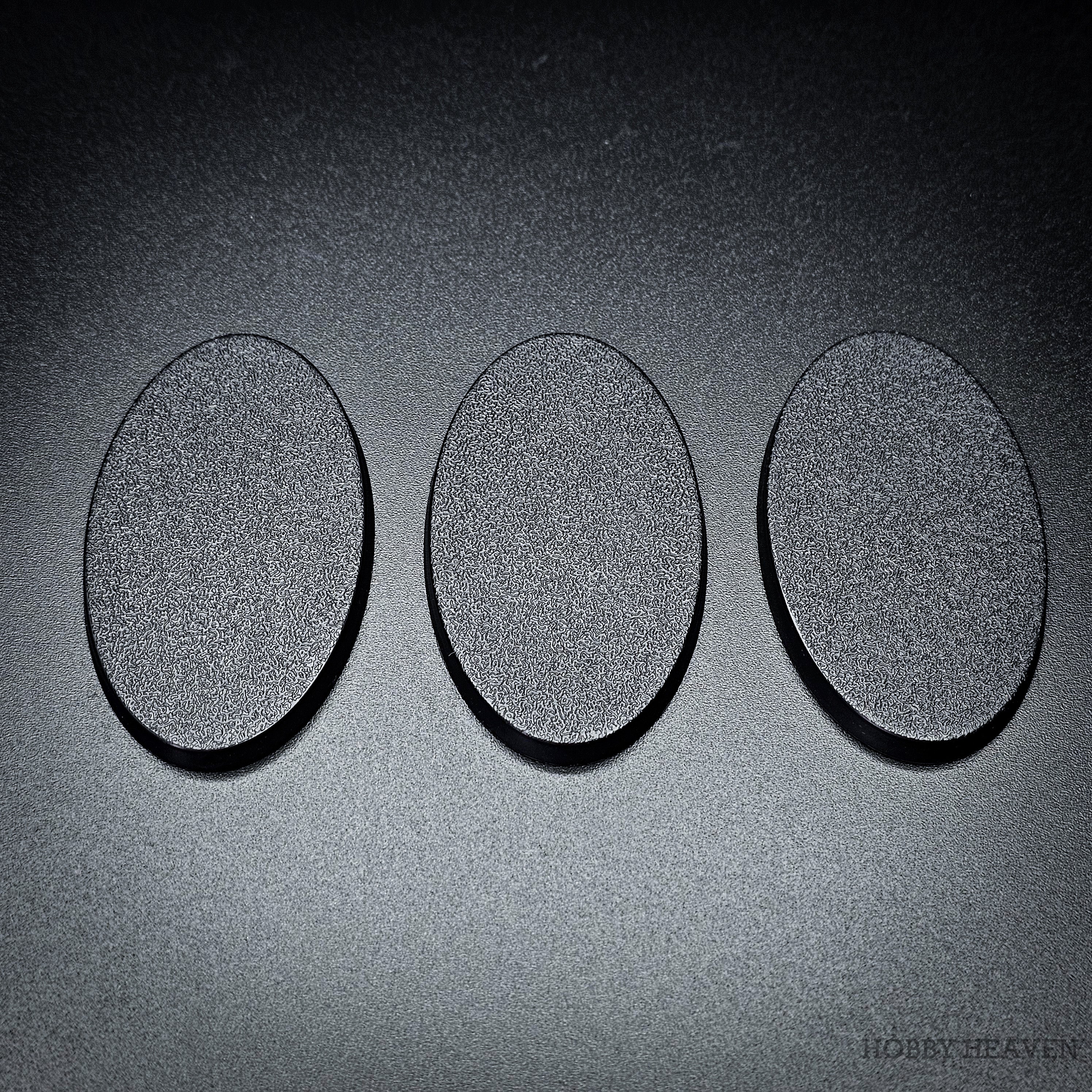 Three 60x35mm oval plain plastic bases in a row, matte black with textured tops facing up