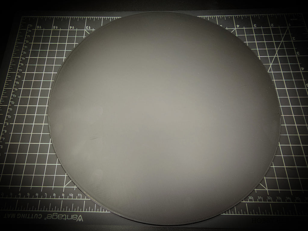 Top-down photograph of the 300mm round smooth PLA base on a cutting mat, showing the clean matte surface with faint concentric FDM layer lines.