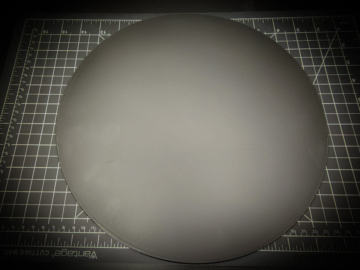 Top-down photograph of the 300mm round smooth PLA base on a cutting mat, showing the clean matte surface with faint concentric FDM layer lines.