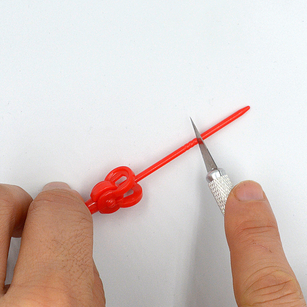 Hands demonstrating how to split AK Interactive holding pin along the pre-scored groove using a hobby knife blade showing the red plastic clip-top pin being separated into two halves