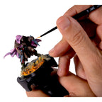 Hand holding AK Interactive Painting Handle while painting a detailed purple and brown fantasy miniature with an AK brush showing the handle in practical use with a fully based and painted model