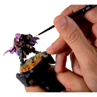 Hand holding AK Interactive Painting Handle while painting a detailed purple and brown fantasy miniature with an AK brush showing the handle in practical use with a fully based and painted model
