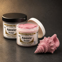 Aves Apoxie Sculpt 1lb Pink — Part A tub open showing pink putty, Part B tub behind, with a detailed sculpted ornate conch shell demonstrating the material's fine detail and smooth finis
