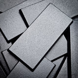 Matte black 50x25mm rectangular plain wargaming bases with a fine textured top, injection-moulded ABS, shown in a pile.
