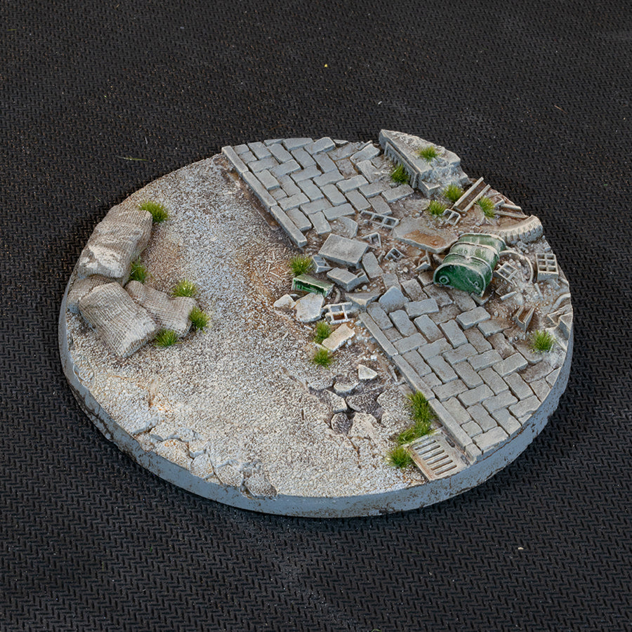 The 100mm base turned again: paving, a toppled green drum and scattered slabs, with grass tufts through the cracks