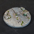 The 100mm base turned again: paving, a toppled green drum and scattered slabs, with grass tufts through the cracks