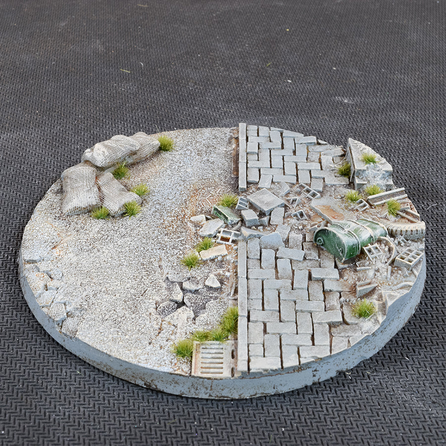 The 100mm base from the side: sandbags on gravel at the left, paving broken by rubble and a street drain at the right