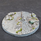 The 100mm base from the side: sandbags on gravel at the left, paving broken by rubble and a street drain at the right