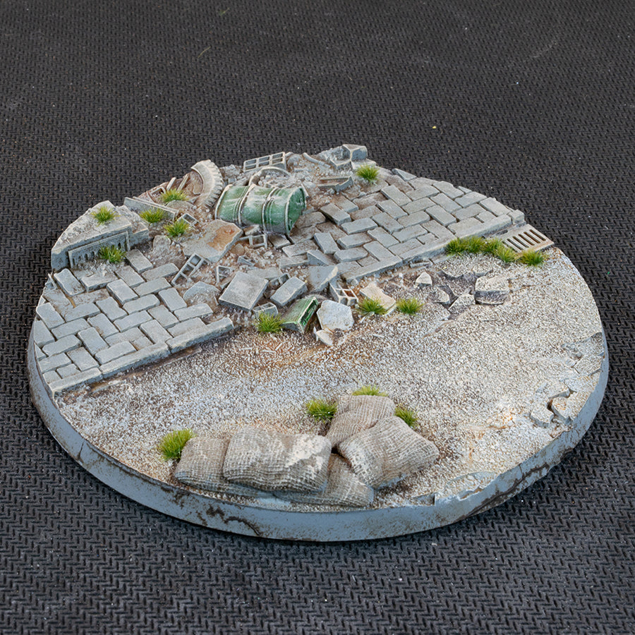 Urban Warfare 100mm base: brick paving and a green fuel drum on its side, with sandbags on open grit in front